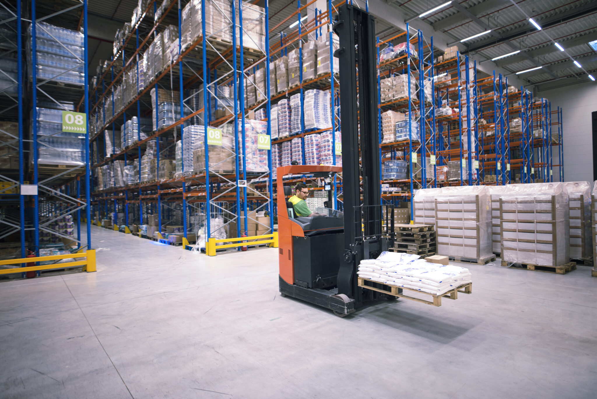 Worker operating forklift machine and relocating goods in large warehouse center.